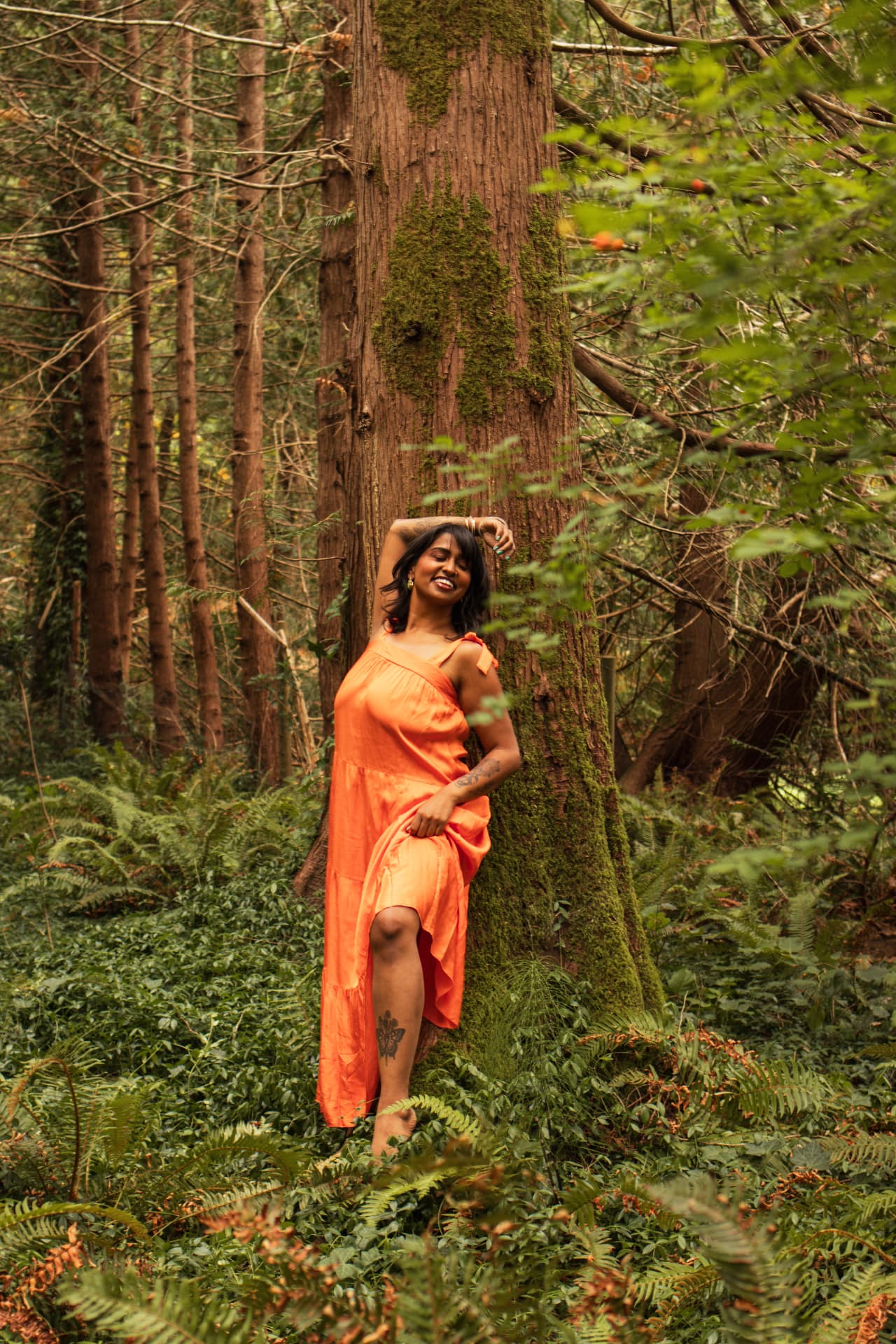 A woman in an orange dress leaning against a big mossy tree